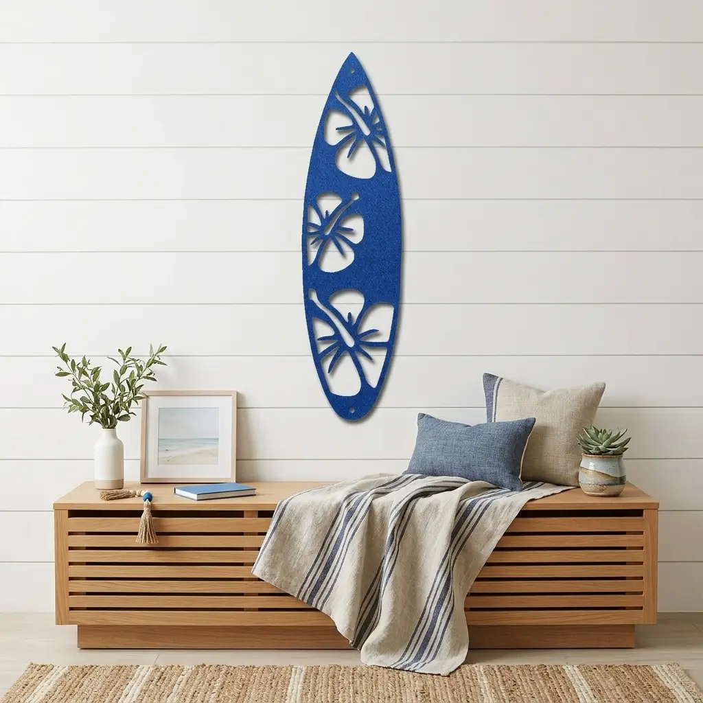 A blue felt wall decor piece shaped like a surfboard with cutout floral designs, hanging on a white shiplap wall above a wooden bench styled with pillows, a throw blanket, and small decor items.